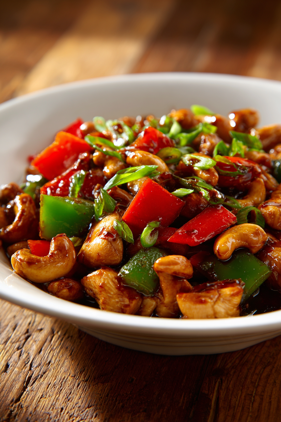 Close-up of authentic cashew chicken stir-fry with diced chicken, crunchy cashews, green bell peppers, and scallions coated in glossy soy and oyster sauce served in a white bowl on rustic wood.