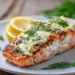 Close-up of a baked salmon fillet topped with creamy garlic herb Boursin cheese spread, garnished with a lemon slice and fresh dill on a white plate with warm natural lighting