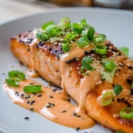 Close-up of crispy pan-seared salmon fillet drizzled with creamy spicy bang bang sauce on white plate, garnished with chopped green onions and sesame seeds