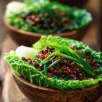 Close-up of rustic cabbage leaf bowls filled with sautéed ground beef and green cabbage, garnished with sliced green onions and red pepper flakes, with a warm sesame oil glaze