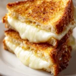 Close-up top-down photo of a grilled mozzarella sandwich cut diagonally, showing crispy golden-brown bread and melted mozzarella cheese oozing on a plain white plate