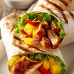 Close-up of Hawaiian BBQ chicken wraps cut in half, stacked on a white plate with bright pineapple chunks, red bell peppers, shredded lettuce, and sweet BBQ sauce in a ramekin.