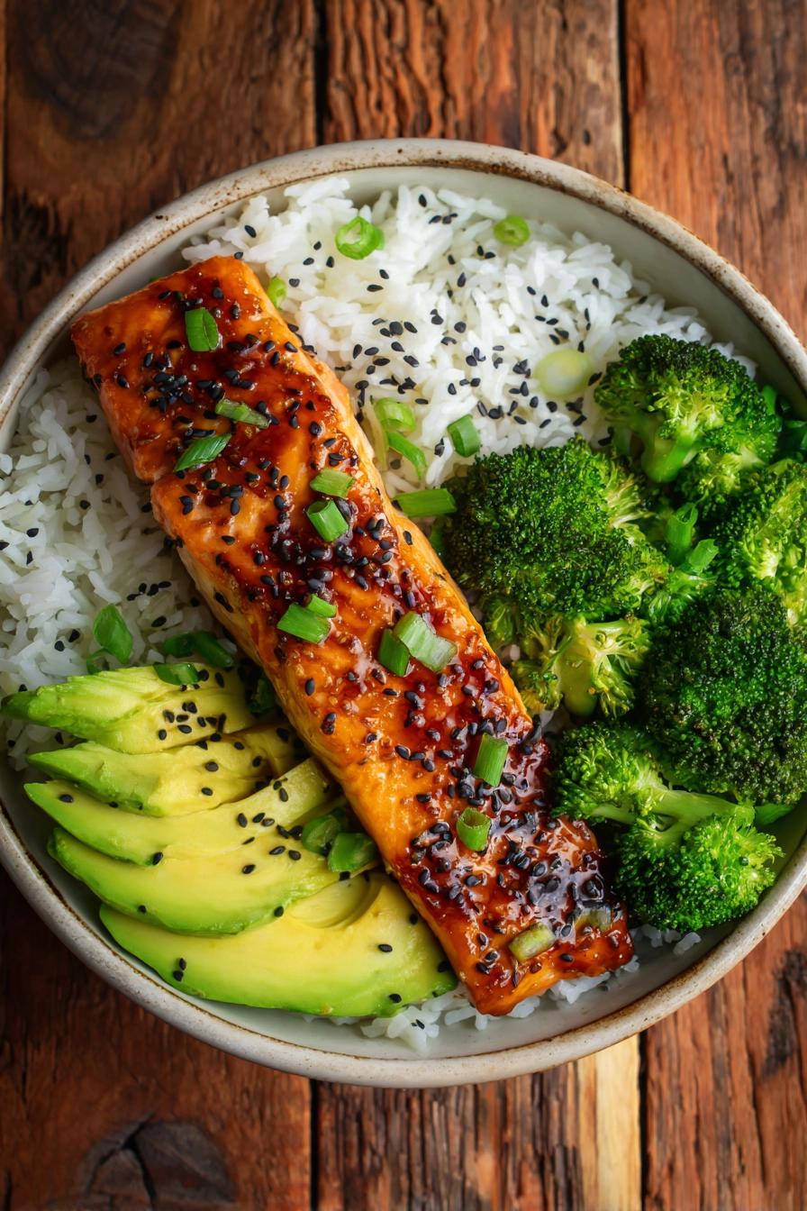 Close-up of honey glazed pan-seared salmon fillet with sesame seeds on fluffy jasmine rice, steamed broccoli florets, and sliced avocado arranged neatly with chopped green onions