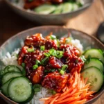 Close-up of Sweet and Spicy Gochujang Chicken Bowl with tender chicken thighs coated in glossy red sauce, white rice, julienned carrots, sliced cucumbers, green onions, and toasted sesame seeds on rustic wooden surface