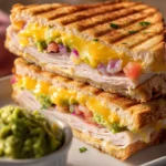 Diagonal cut Turkey Guacamole Grilled Cheese Sandwich stacked on a white rectangular plate, showing melted cheddar, sliced turkey, and fresh green guacamole with a ramekin of extra guacamole on rustic wooden surface