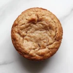 2 ingredient protein muffin - the image is a close-up of a round, golden-brown muffin on a white marble surface. the muffin appears to be freshly baked and has a crumbly texture. it is slightly tilted to the side, with the top half of the muffin slightly overlapping the bottom half. the background is blurred, making the muffins the focal point of the image.
