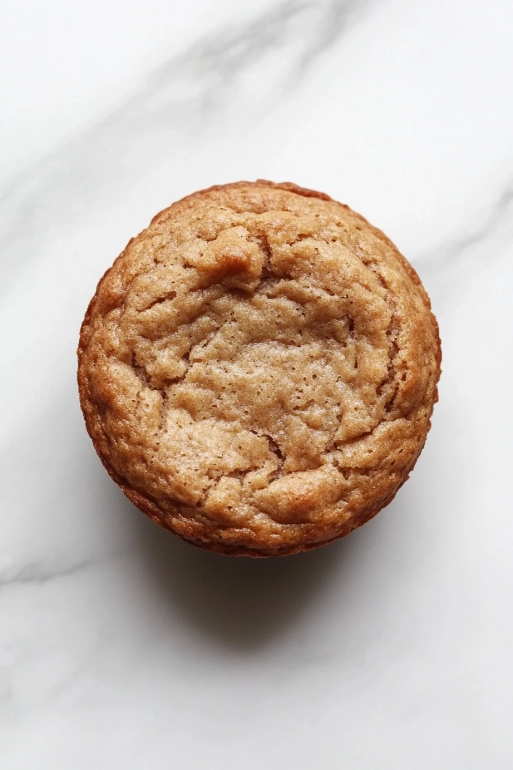 2 ingredient protein muffin - the image is a close-up of a round, golden-brown muffin on a white marble surface. the muffin appears to be freshly baked and has a crumbly texture. it is slightly tilted to the side, with the top half of the muffin slightly overlapping the bottom half. the background is blurred, making the muffins the focal point of the image.