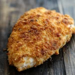 Air fryer chicken breast crispy - the image shows a piece of breaded chicken breast on a wooden surface. the chicken breast is golden brown and appears to be seasoned with herbs and spices. the breadcrumbs are scattered across the top of the chicken breast, giving it a crispy texture. the wooden surface has a weathered and aged appearance, with visible cracks and chips. the background is blurred, making the chicken the focal point of the image.