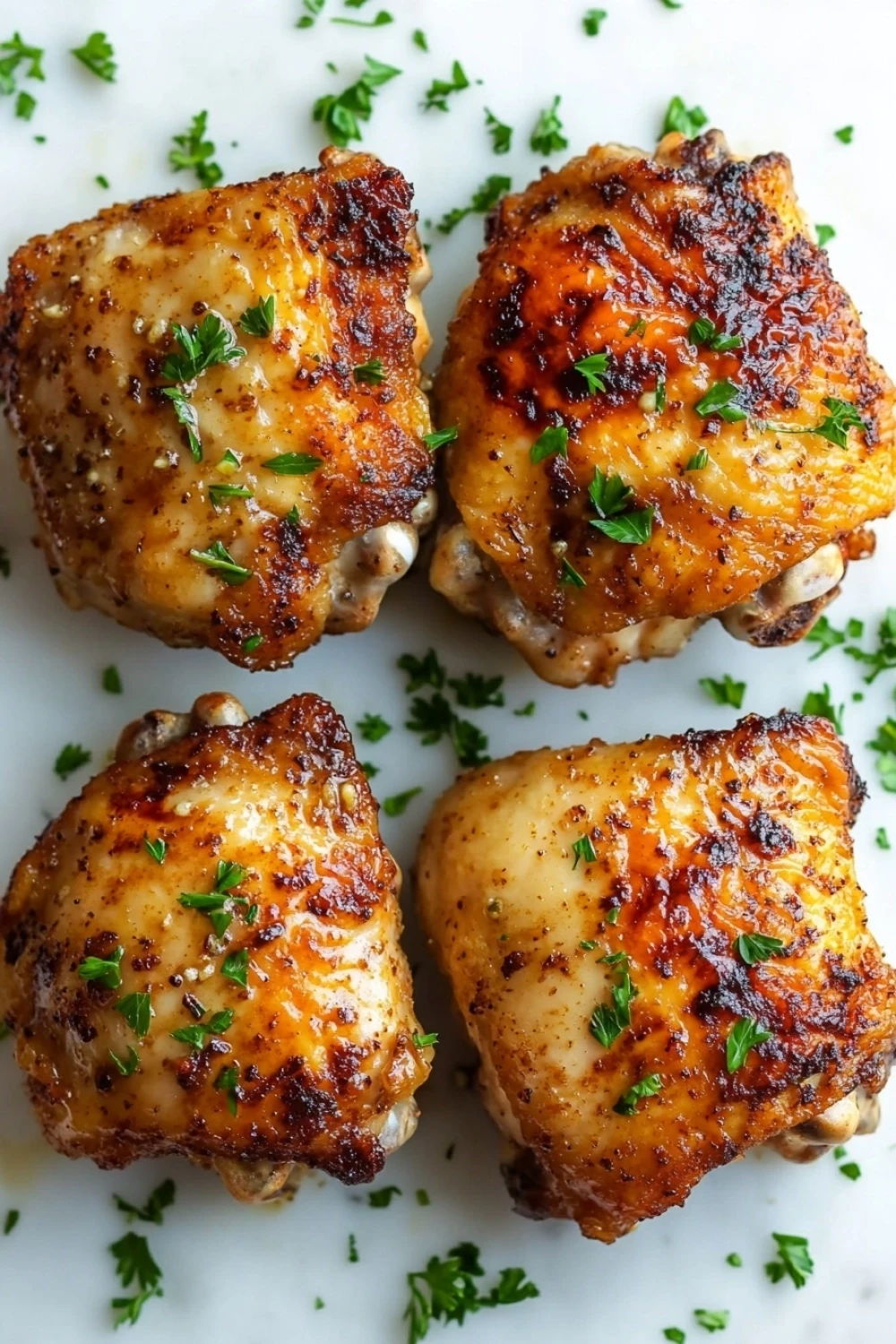 Air fryer garlic butter chicken thigh - the image shows four pieces of grilled chicken thighs on a white plate. the chicken thighs are golden brown and appear to be seasoned with herbs and spices. the plate is garnished with a sprig of parsley, adding a pop of green color to the dish. the background is blurred, making the chicken thighs the focal point of the image.