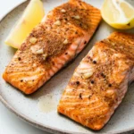 Air fryer salmon cook time - the image shows two pieces of salmon on a gray plate. the salmon is cooked to a golden brown color and appears to be seasoned with herbs and spices. there are two lemon wedges on the plate, one on each side of the salmon. the plate is sitting on a white marble countertop. the background is blurred, but it seems to be a kitchen countertop with a gray and white striped napkin.