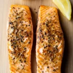 Air fryer salmon crispy - the image shows two pieces of salmon on a wooden cutting board. the salmon is cooked to a golden brown color and appears to be seasoned with black pepper flakes. there are two lemon wedges on either side of the salmon, one on top of the other. the background is a light-colored wood surface.