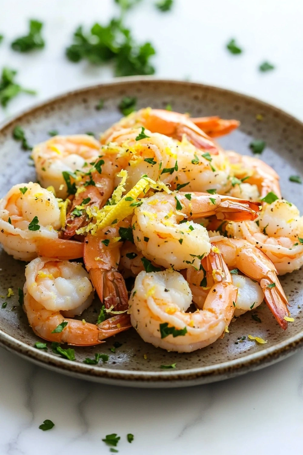 Air fryer shrimp lemon - the image shows a plate of cooked shrimp on a white marble countertop. the shrimp are arranged in a circular pattern on the plate, with some overlapping each other. they are orange in color and appear to be seasoned with herbs and spices. the plate is garnished with chopped parsley, adding a pop of green color to the dish. the background is blurred, but it appears to be a kitchen countertop with more parsley scattered around.