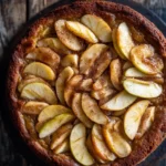 Apple cake korean - the image is a close-up of a freshly baked apple tart on a wooden table. the tart has a golden brown crust and is topped with sliced apples arranged in a circular pattern. the apples are cut into thin slices and are arranged in an overlapping manner, with some overlapping each other. the top of the tart is drizzled with a caramel sauce, giving it a shiny appearance. the table has a rustic, weathered look, with visible grain and knots. the overall mood of the image is warm and inviting.