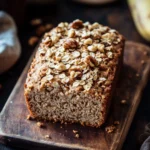 Banana bread no baking - the image shows a freshly baked loaf of bread on a wooden cutting board. the bread is golden brown in color and has a crumbly texture. it is topped with chopped nuts, giving it a crunchy appearance. in the background, there is a banana, a cup of coffee, and a few scattered nuts on the table. the overall mood of the image is rustic and cozy.
