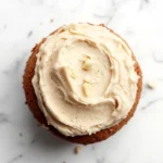 Banana cake frosting - the image is a close-up of a cupcake on a white marble surface. the cupcake is round and has a golden brown crust. it is topped with a generous layer of creamy white frosting that has a swirl pattern on top. there are small pieces of nuts scattered around the cupcake, adding a crunchy texture to the overall appearance. the frosting appears to be smooth and creamy, with a light dusting of powdered sugar on top for added flavor.