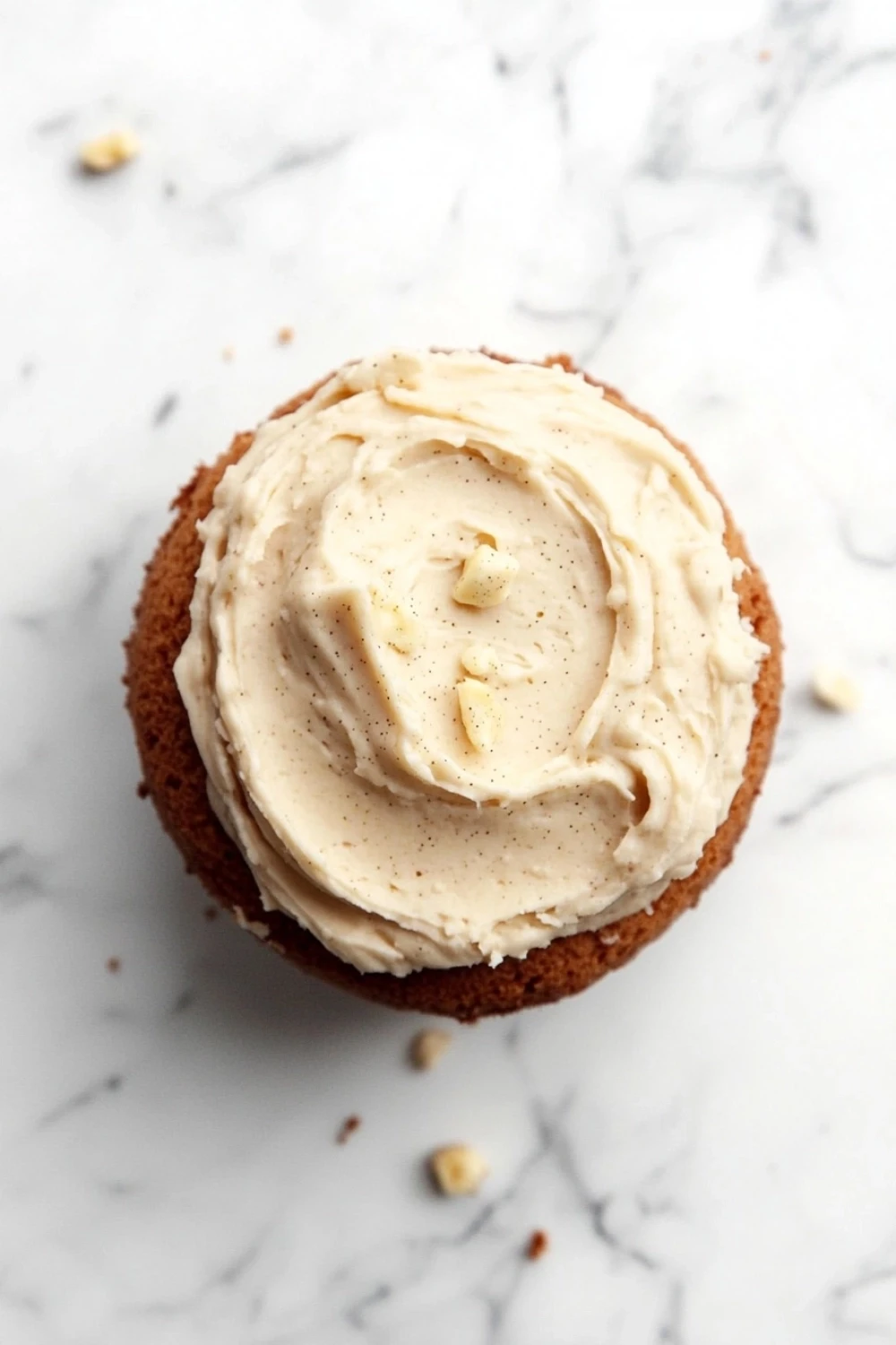 Banana cake frosting - the image is a close-up of a cupcake on a white marble surface. the cupcake is round and has a golden brown crust. it is topped with a generous layer of creamy white frosting that has a swirl pattern on top. there are small pieces of nuts scattered around the cupcake, adding a crunchy texture to the overall appearance. the frosting appears to be smooth and creamy, with a light dusting of powdered sugar on top for added flavor.