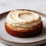 Banana cake sticker - the image shows a round cake on a white plate. the cake appears to be a bundt cake with a golden brown crust and a creamy white frosting on top. the frosting is swirled in a circular pattern and has a smooth texture. the plate is round and has an orange rim. the background is a plain white surface.