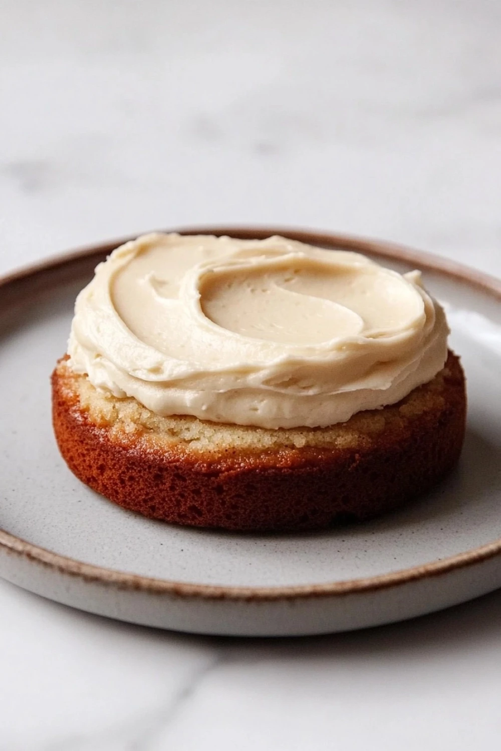 Banana cake sticker - the image shows a round cake on a white plate. the cake appears to be a bundt cake with a golden brown crust and a creamy white frosting on top. the frosting is swirled in a circular pattern and has a smooth texture. the plate is round and has an orange rim. the background is a plain white surface.