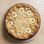 Banana cake vegan gluten free - the image is of a round cake on a white cake stand. the cake appears to be freshly baked and has a golden brown crust on top. on top of the cake, there are several sliced bananas arranged in a circular pattern. the bananas are evenly spaced and appear to be ripe and juicy. the background is a light-colored wooden surface.