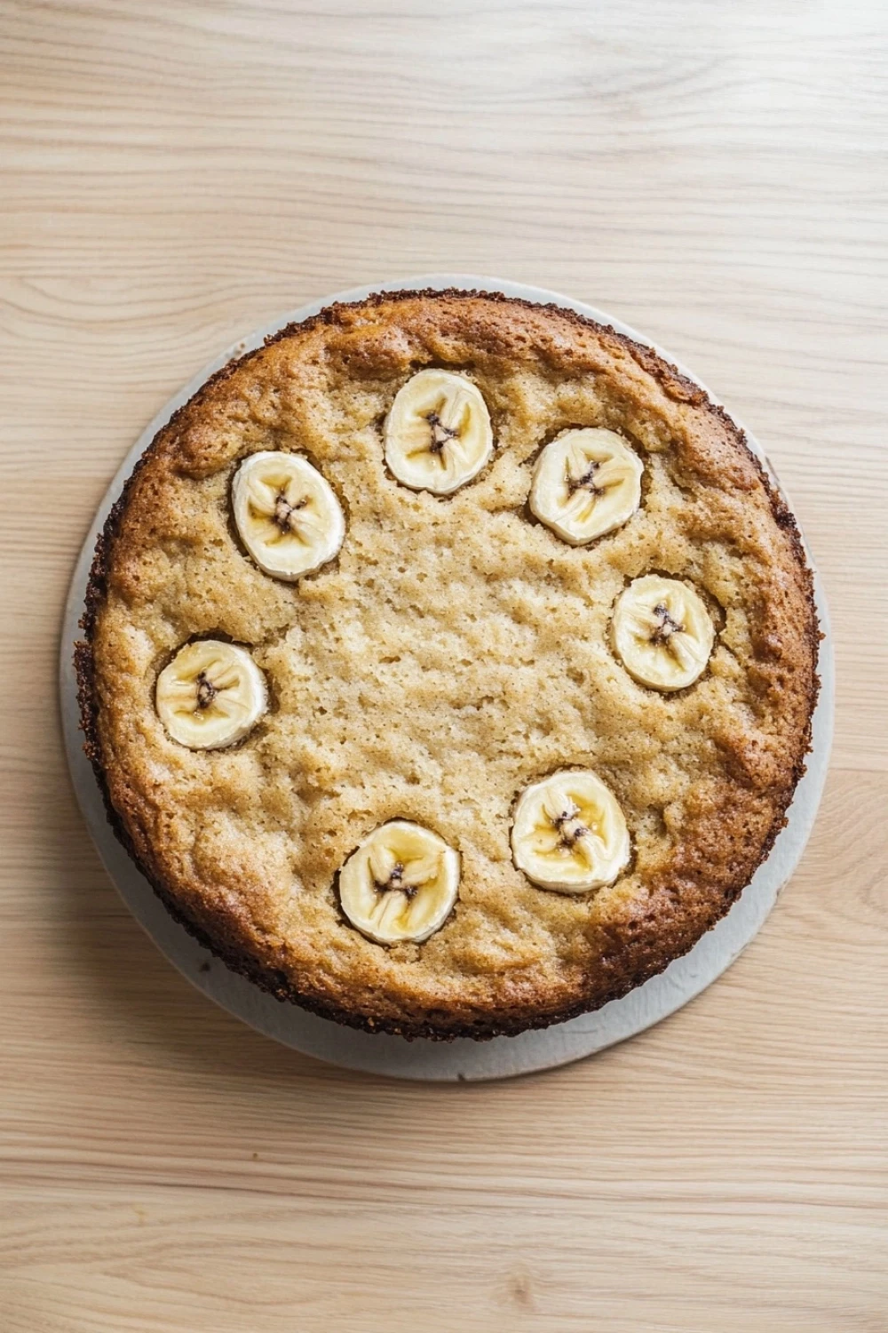 Banana cake vegan gluten free - the image is of a round cake on a white cake stand. the cake appears to be freshly baked and has a golden brown crust on top. on top of the cake, there are several sliced bananas arranged in a circular pattern. the bananas are evenly spaced and appear to be ripe and juicy. the background is a light-colored wooden surface.
