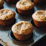 Banana muffin almond flour - the image shows a muffin tin filled with freshly baked muffins. the muffins are golden brown and have a crumbly texture. they are arranged in a single layer in the tin, with some overlapping each other. the tin is placed on a wooden table, and the background is blurred, making the muffins the focal point of the image.