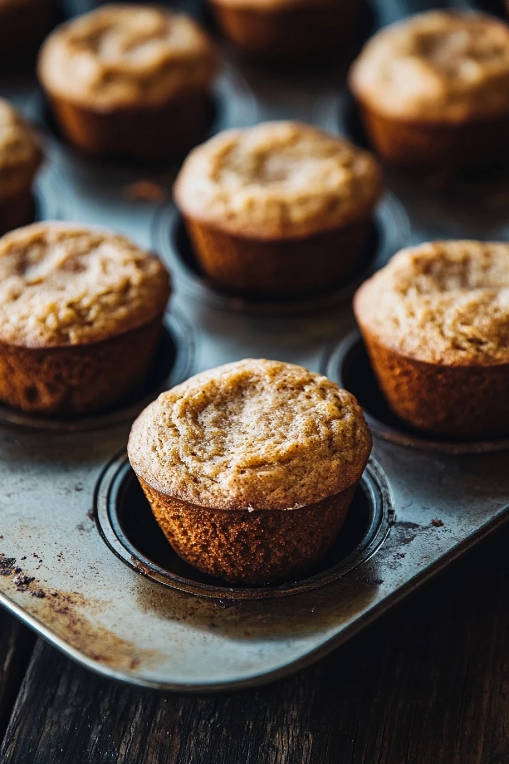 Banana muffin almond flour - the image shows a muffin tin filled with freshly baked muffins. the muffins are golden brown and have a crumbly texture. they are arranged in a single layer in the tin, with some overlapping each other. the tin is placed on a wooden table, and the background is blurred, making the muffins the focal point of the image.