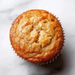 Banana muffin maple syrup - the image is a close-up of a freshly baked muffin on a white marble surface. the muffin is golden brown in color and has a crumbly texture. it is covered in a light dusting of powdered sugar, giving it a shiny appearance. the edges of the muffin are slightly curled, indicating that it has been freshly baked. the background is blurred, making the muffins the focal point of the image.
