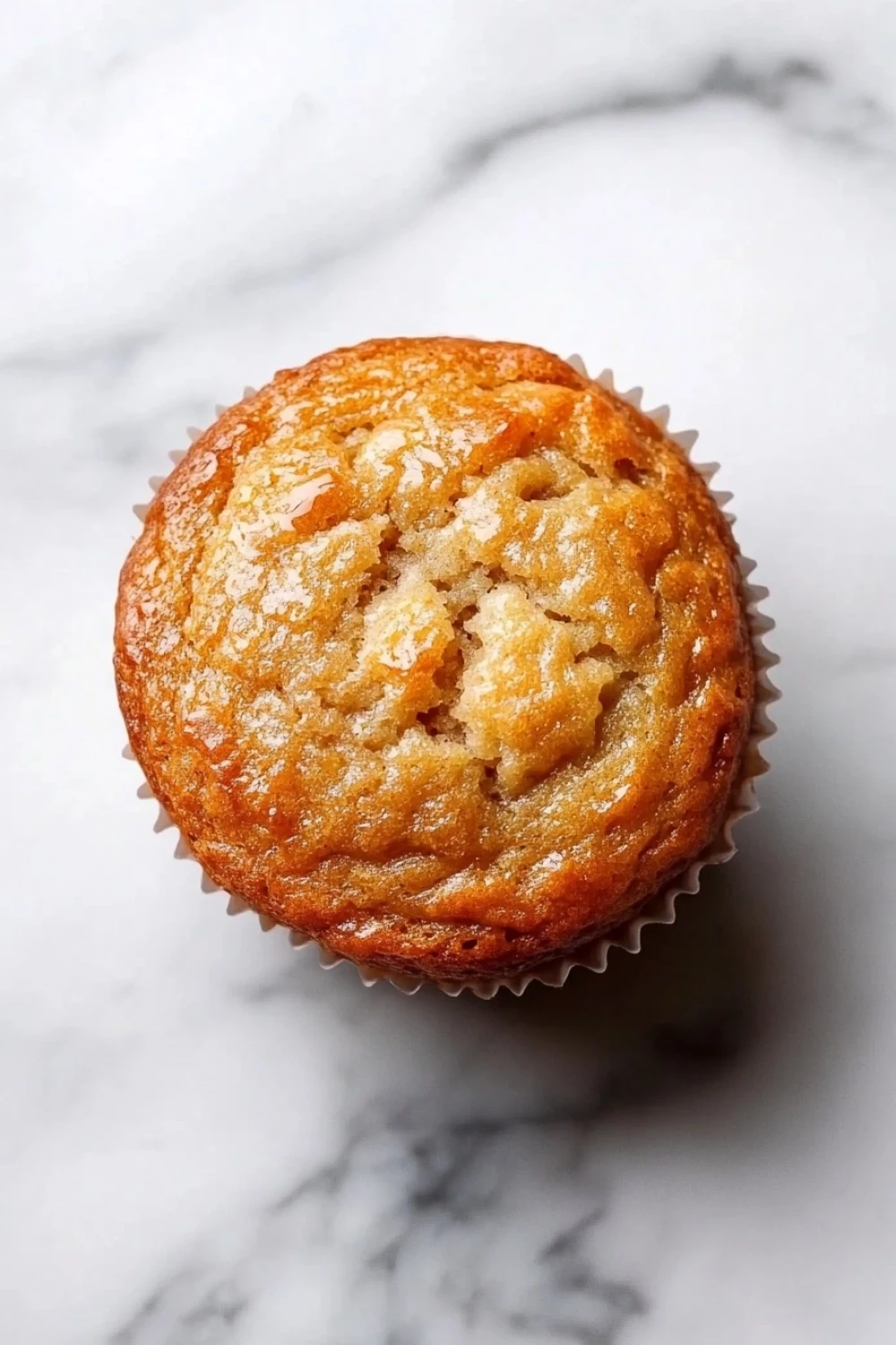 Banana muffin maple syrup - the image is a close-up of a freshly baked muffin on a white marble surface. the muffin is golden brown in color and has a crumbly texture. it is covered in a light dusting of powdered sugar, giving it a shiny appearance. the edges of the muffin are slightly curled, indicating that it has been freshly baked. the background is blurred, making the muffins the focal point of the image.