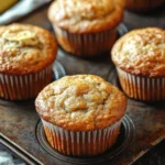 Banana muffin with sour cream - the image shows a group of freshly baked banana muffins on a metal tray. the muffins are golden brown in color and have a crumbly texture. they are arranged in a row on the tray, with a few bananas visible in the background. the tray is placed on a wooden table with a gray cloth napkin on the side. the overall mood of the image is warm and inviting.