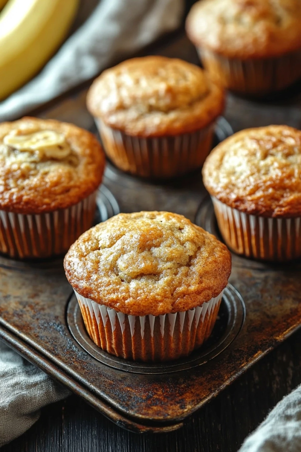 Banana muffin with sour cream - the image shows a group of freshly baked banana muffins on a metal tray. the muffins are golden brown in color and have a crumbly texture. they are arranged in a row on the tray, with a few bananas visible in the background. the tray is placed on a wooden table with a gray cloth napkin on the side. the overall mood of the image is warm and inviting.