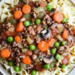 Beef casserole with ground beef and slow cooker - the image is a close-up of a bowl of beef noodle soup. the soup is made with ground beef, carrots, peas, onions, and mushrooms. the noodles are long and thin, and the soup is a rich, dark brown color. the vegetables are bright orange and appear to be fresh. the bowl is white and is sitting on a wooden table. the background is blurred, but it appears to be a kitchen countertop.