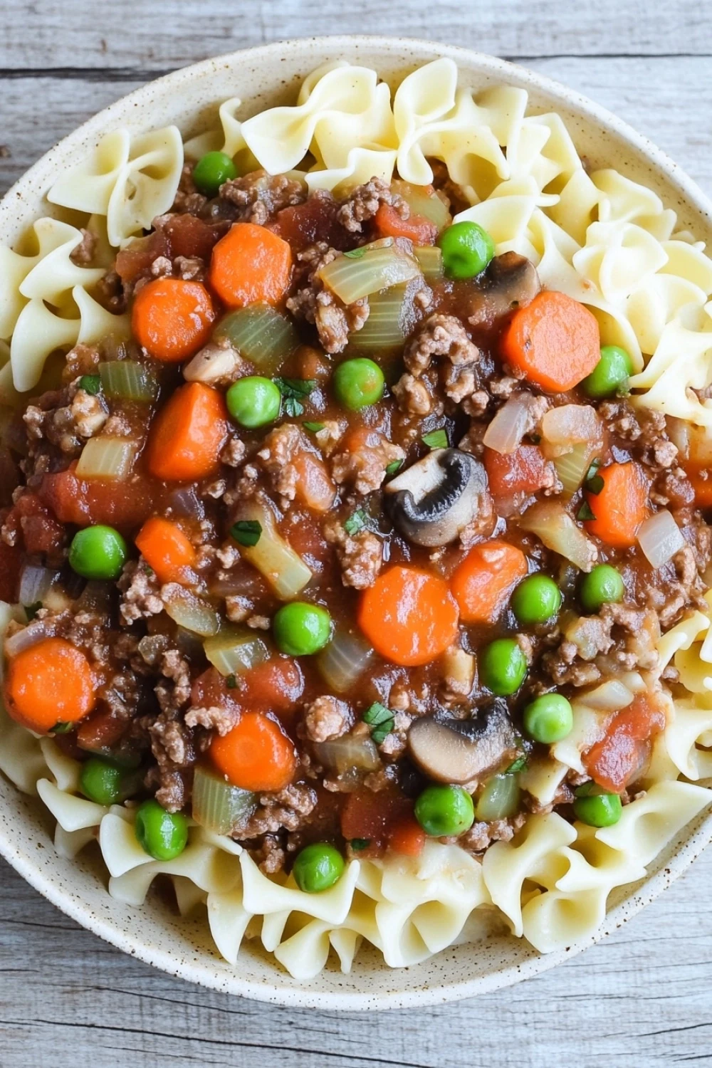 Beef casserole with ground beef and slow cooker - the image is a close-up of a bowl of beef noodle soup. the soup is made with ground beef, carrots, peas, onions, and mushrooms. the noodles are long and thin, and the soup is a rich, dark brown color. the vegetables are bright orange and appear to be fresh. the bowl is white and is sitting on a wooden table. the background is blurred, but it appears to be a kitchen countertop.