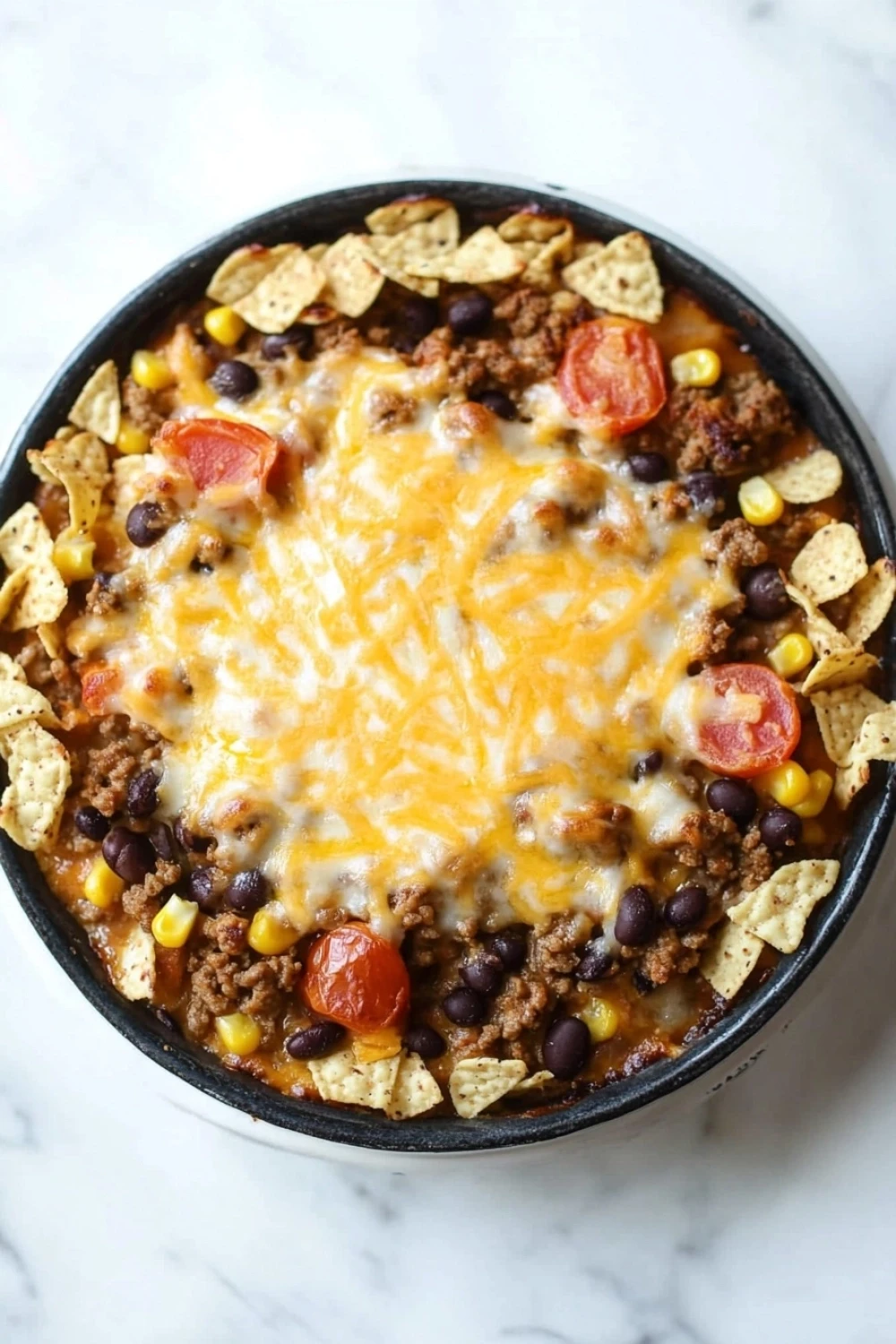 Beef casserole with ground beef and tortilla chips - the image is of a bowl of chili con carne. the chili is made with ground beef, black beans, corn kernels, and diced tomatoes. it is topped with melted cheese and tortilla chips. the bowl is black and is sitting on a white marble countertop.