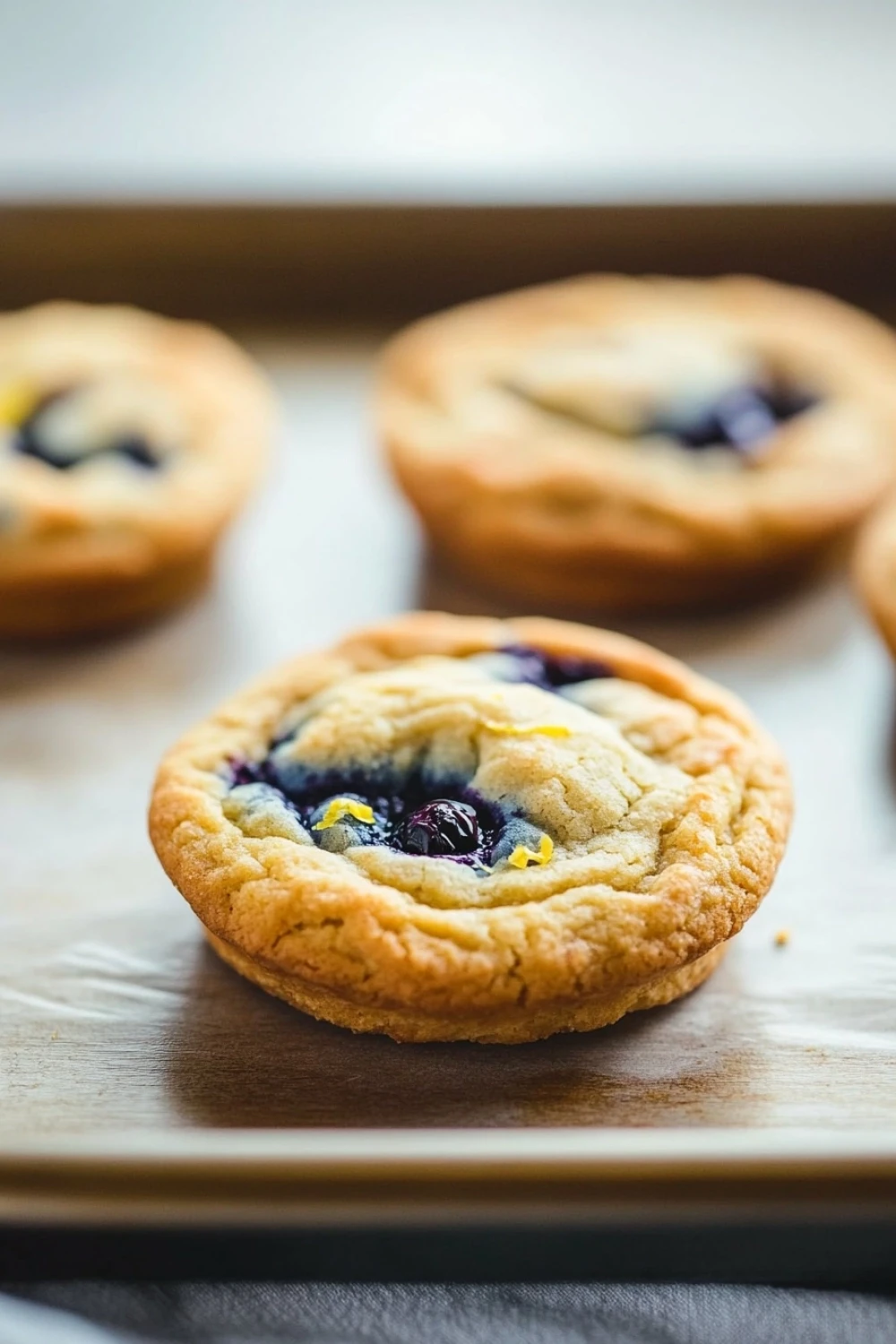 Blueberry lemon muffin top cookies - the image shows a row of freshly baked blueberry tarts on a wooden baking tray. the tarts are golden brown in color and have a crumbly texture. they are filled with blueberries and yellow zest, which are visible in the center of each tarts. the tray is lined with parchment paper, and the background is blurred, making the tarts the focal point of the image.