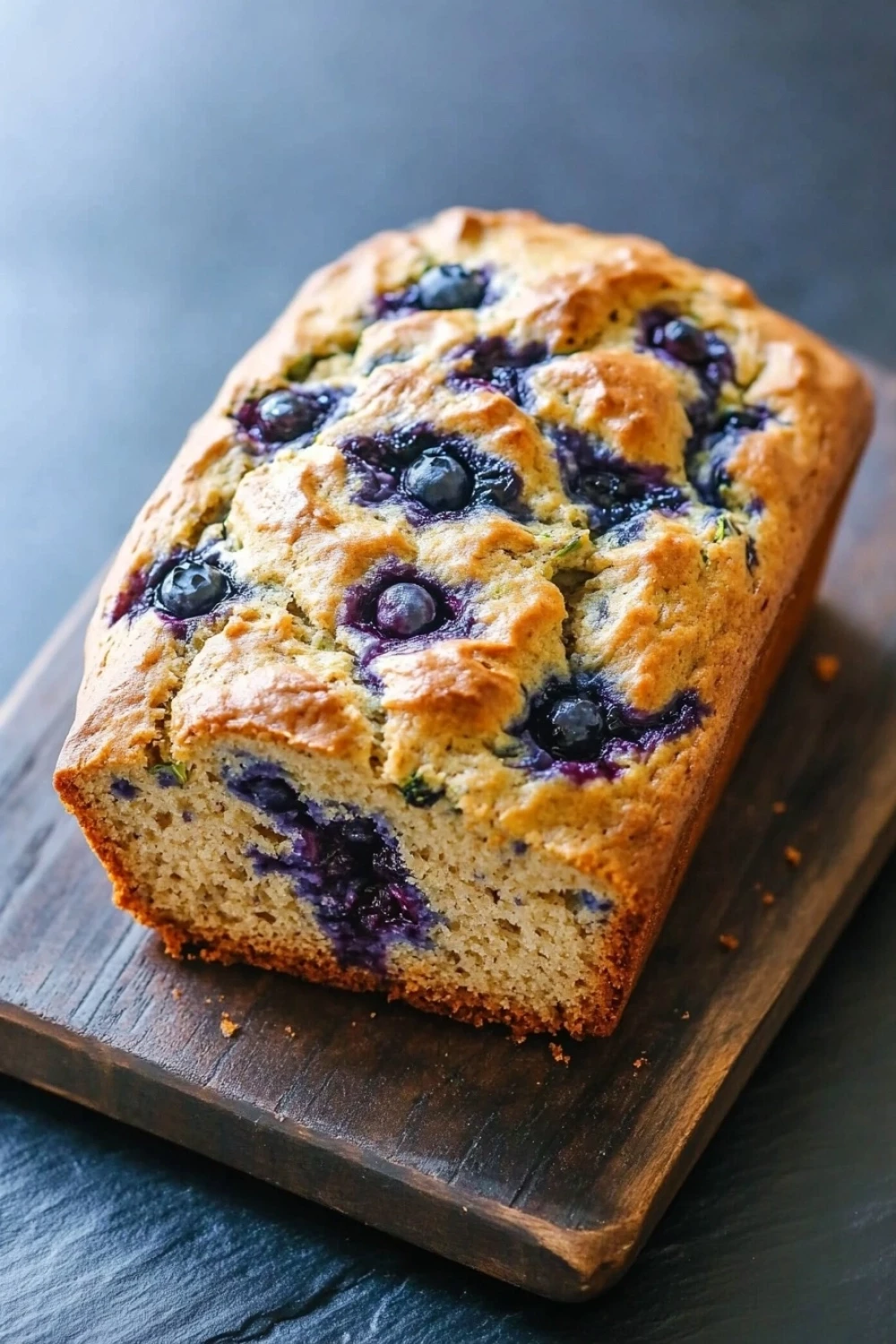 Blueberry zucchini bread - the image shows a freshly baked loaf of blueberry bread on a wooden cutting board. the bread has a golden brown crust and is topped with fresh blueberries. the blueberries are arranged in a circular pattern on top of the bread, with some overlapping each other. the cutting board is placed on a dark grey countertop. the background is blurred, making the bread the focal point of the image.