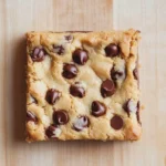 Chewy cake mix cookie bars - the image is a close-up of a square-shaped cookie bar on a wooden surface. the cookie bar is golden brown in color and has a crumbly texture. it is topped with chocolate chips, giving it a crunchy appearance. the chocolate chips are scattered across the top of the cookie bar, adding a pop of color to the dish. the background is a light beige color, which contrasts with the warm tones of the wood.