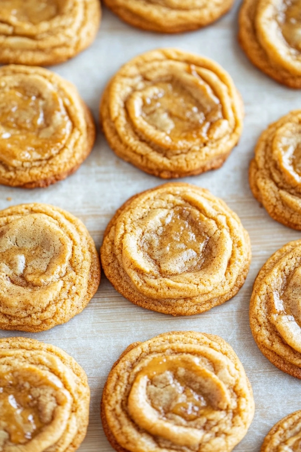 Chewy cookie butter cookies - the image shows a tray of freshly baked cinnamon rolls on a baking sheet. the rolls are golden brown in color and have a swirl pattern on top. they are dusted with a light dusting of powdered sugar, giving them a shiny appearance. the tray is lined with parchment paper, and the rolls are arranged in a neat row. the background is blurred, but it appears to be a kitchen countertop.