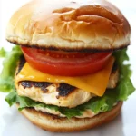 Chicken breast burgers - the image is of a burger on a white marble countertop. the burger is made with a toasted bun and is topped with a generous helping of melted cheese, a slice of tomato, and a bed of lettuce. the cheese is a bright yellow color and appears to be melted and oozing out of the sides. the tomato is a deep red color and is placed on top of the cheese. the lettuce is a light green color and looks fresh and wilted. the background is blurred, making the burger the focal point of the image.