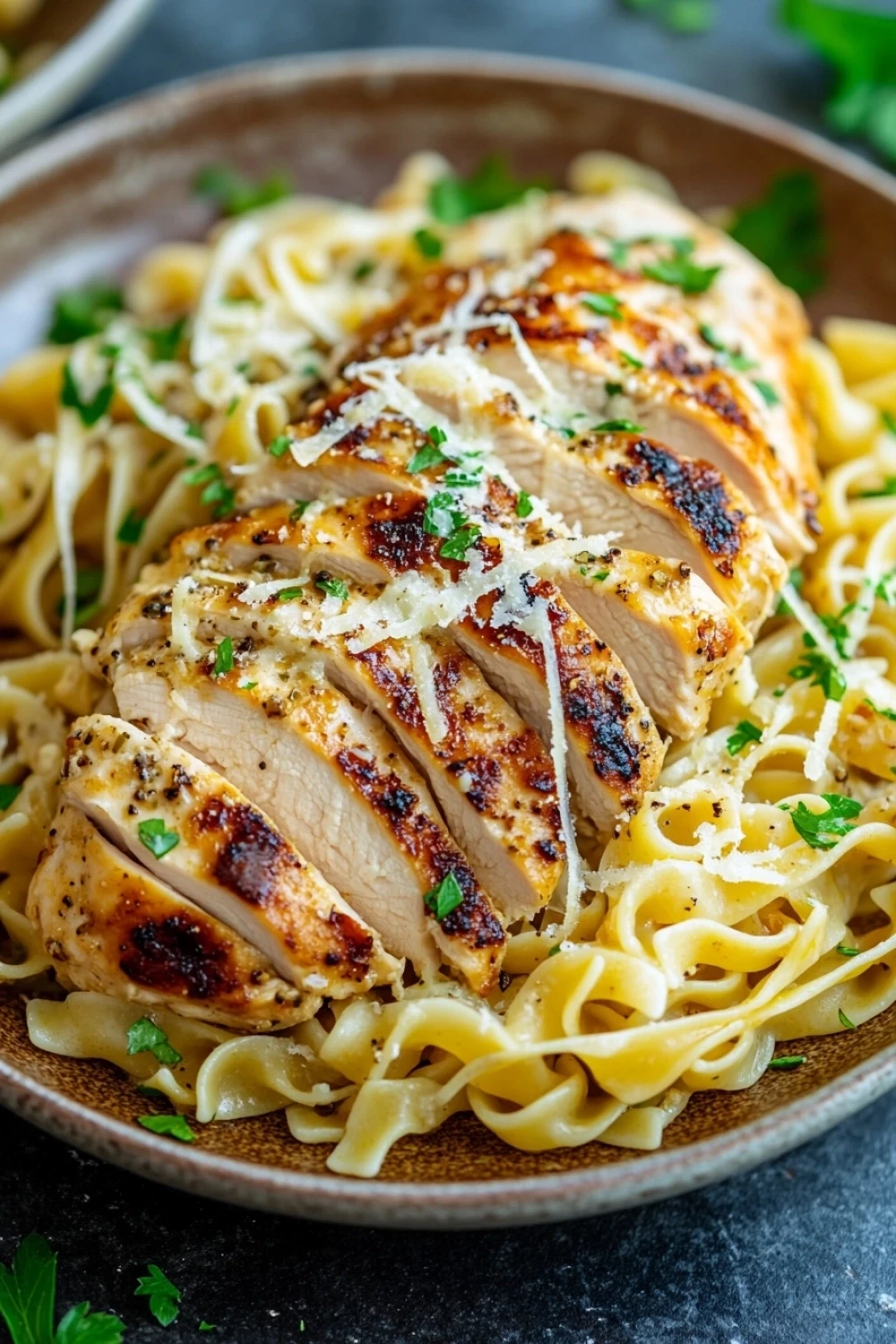 Chicken breast dinner noodles - the image shows a plate of fettuccine alfredo pasta with grilled chicken. the pasta is cooked al dente and is topped with grated parmesan cheese. the chicken is cooked to a golden brown color and appears to be seasoned with herbs and spices. the dish is garnished with fresh parsley, adding a pop of green color to the dish. the plate is sitting on a dark grey countertop with a few sprigs of parsley scattered around.
