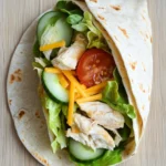 Chicken breast wraps - the image shows a wrap on a wooden table. the wrap is made of tortilla and is filled with a variety of ingredients. on top of the wrap, there are shredded chicken, sliced cucumbers, sliced tomatoes, and shredded cheese. the chicken appears to be seasoned with herbs and spices, and the vegetables are arranged in a colorful and appetizing manner.