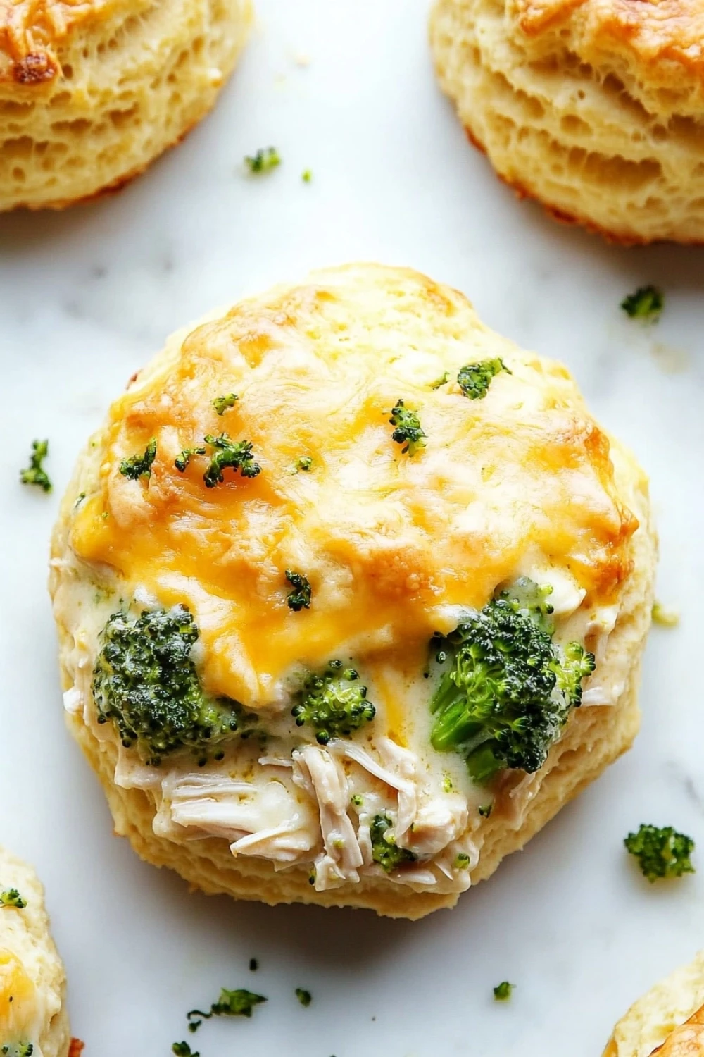 Chicken broccoli casserole biscuits - the image shows a close-up of a round, golden-brown biscuit with a layer of melted cheese on top. the cheese is melted and bubbly, and there are small pieces of broccoli scattered around the biscuit. the biscuit appears to be freshly baked and is sitting on a white marble surface. there are a few more biscuit rolls visible in the background, but they are slightly blurred. the overall color scheme of the image is warm and inviting.