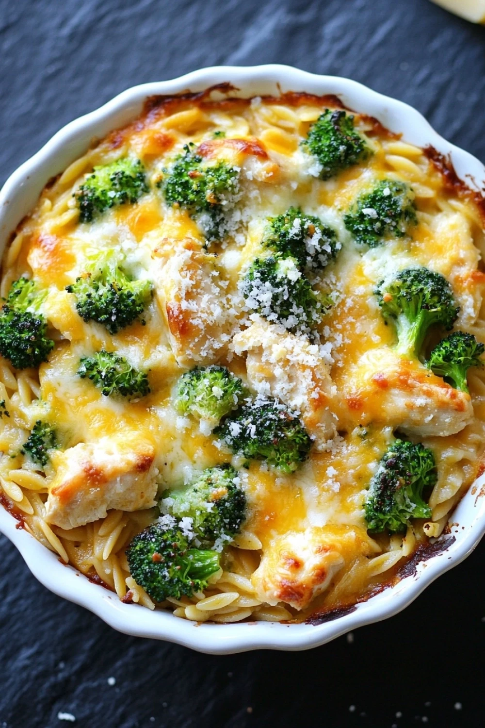 Chicken broccoli casserole with orzo - the image is of a casserole dish filled with a creamy pasta dish. the dish appears to be made with broccoli and cheese, and it is topped with grated parmesan cheese. the pasta is long and thin, and the dish is sitting on a black countertop. the broccoli is bright green and looks fresh and healthy. the cheese is melted and bubbly, and there are small pieces of broccoli scattered throughout the dish.