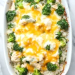 Chicken broccoli rice casserole - the image shows a white rectangular baking dish filled with a casserole dish. the dish is filled with white rice and topped with a layer of melted cheese. the cheese is a bright yellow color and appears to be melted and bubbly. on top of the cheese, there are small pieces of broccoli florets scattered throughout the dish. there are also a few pieces of chicken visible in the dish, which are also covered in the cheese. a white cloth napkin is visible on the right side of the image. the background is a white marble countertop.