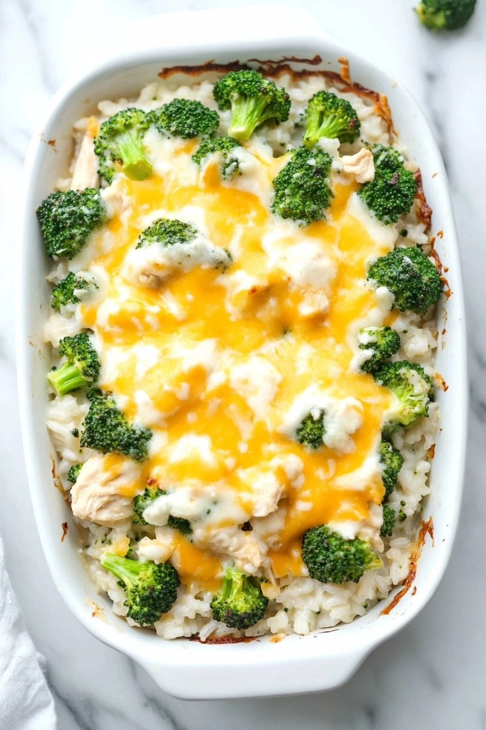 Chicken broccoli rice casserole - the image shows a white rectangular baking dish filled with a casserole dish. the dish is filled with white rice and topped with a layer of melted cheese. the cheese is a bright yellow color and appears to be melted and bubbly. on top of the cheese, there are small pieces of broccoli florets scattered throughout the dish. there are also a few pieces of chicken visible in the dish, which are also covered in the cheese. a white cloth napkin is visible on the right side of the image. the background is a white marble countertop.