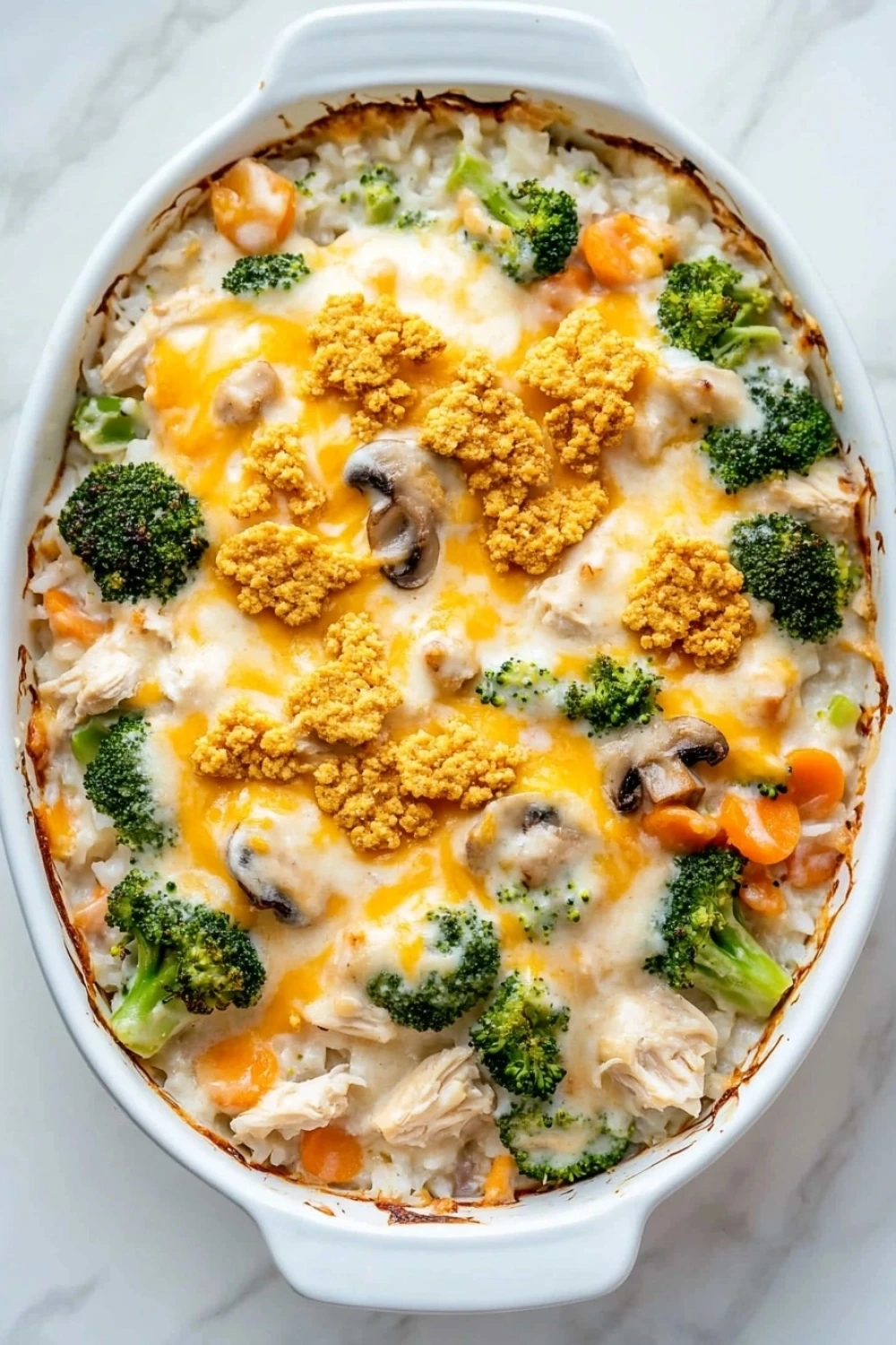 Chicken casserole for a crowd - the image is of a casserole dish filled with a creamy, cheesy dish. the dish appears to be made with chicken, broccoli, carrots, and other vegetables, and is topped with a layer of melted cheese. the cheese is golden brown and bubbly, and there are small pieces of breadcrumbs scattered throughout the dish, giving it a crunchy texture. the casserolle is white and is sitting on a white marble countertop.