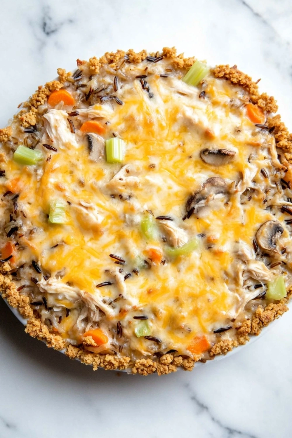 Chicken wild rice casserole - the image is of a freshly baked chicken pot pie on a white marble countertop. the pie has a golden brown crust on top and is filled with a mixture of chicken, mushrooms, carrots, celery, and other vegetables. the cheese is melted and bubbly, and there are small bits of herbs scattered throughout the dish. the dish appears to be freshly baked and ready to eat.