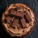 Chocolate cake kitkat - the image is a close-up of a chocolate cake with a layer of chocolate frosting on top. the frosting is a rich, dark brown color and appears to be smooth and creamy. on top of the frosting, there are several small pieces of chocolate arranged in a random pattern. the chocolate bars are dark brown and appear to be freshly made. the cake is placed on a dark, textured surface, possibly a table or countertop. the overall mood of the image is decadent and indulgent.