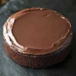 Chocolate cake low sugar - the image is a close-up of a round chocolate cake on a black slate plate. the cake appears to be freshly baked and has a smooth, glossy surface. the top of the cake is covered in a thick layer of chocolate frosting, which is spread evenly across the cake. the frosting is a rich, dark brown color and looks smooth and shiny. the plate is placed on a dark, textured surface, and the background is blurred, making the cake the focal point of the image.