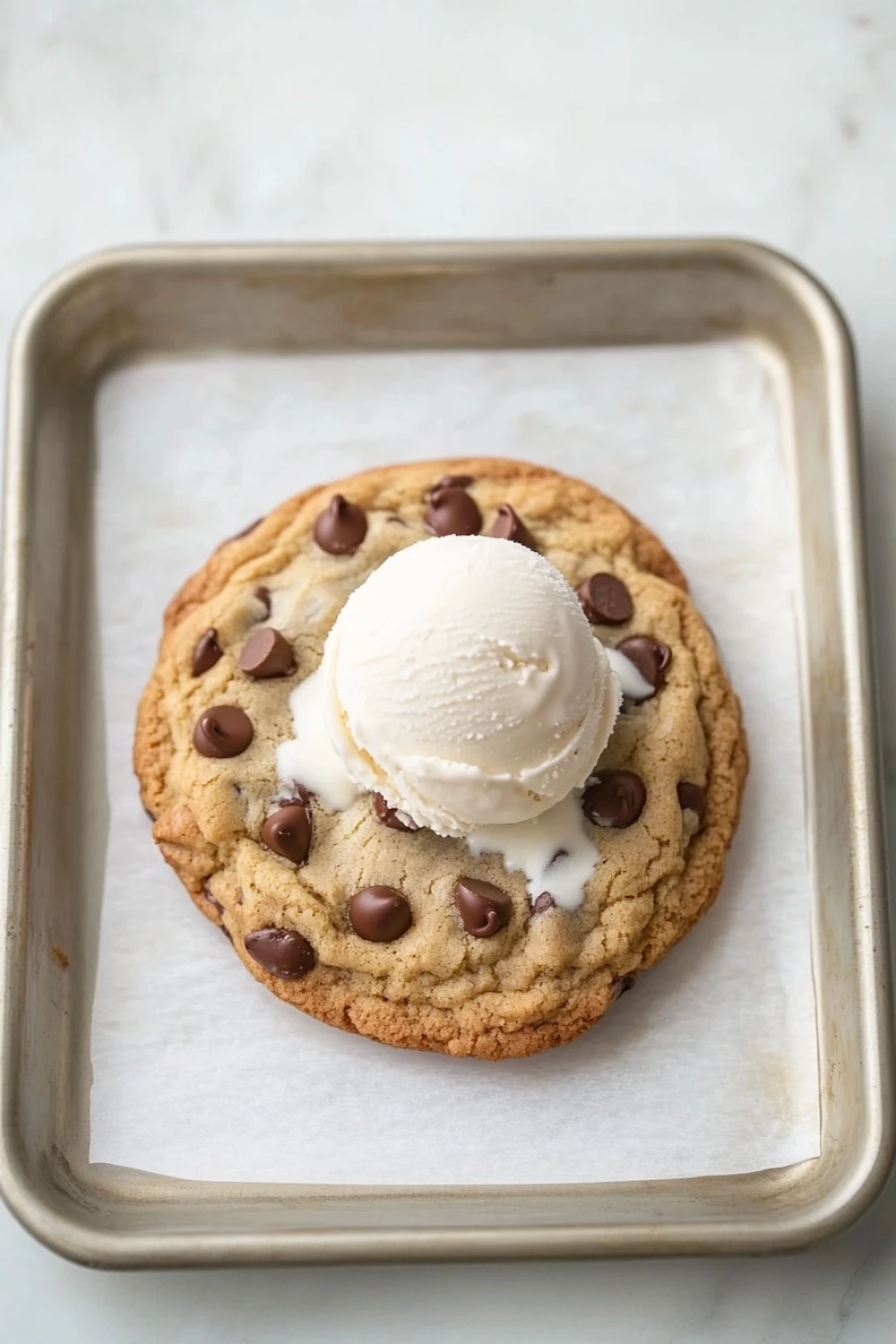 Chocolate chip cookie and ice cream - the image shows a freshly baked chocolate chip cookie on a baking tray. the cookie is golden brown and has chocolate chips scattered throughout. on top of the cookie, there is a scoop of vanilla ice cream. the tray is lined with parchment paper. the background is a white marble countertop.