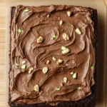 Chocolate zucchini sheet cake - the image is of a square-shaped chocolate cake on a wooden surface. the cake appears to be freshly baked and has a rich, dark brown color. the top of the cake is covered in a thick layer of chocolate frosting, which is swirled in a swirl pattern. the frosting is drizzled over the cake, creating a smooth and glossy texture. there are also small pieces of chopped nuts scattered throughout the frosting. the tray is lined with parchment paper, and the cake looks moist and fluffy.